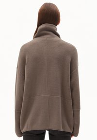 Une personne aux longs cheveux bruns porte une veste en polaire marron surdimensionnée avec un col haut, vue de dos contre un fond uni.