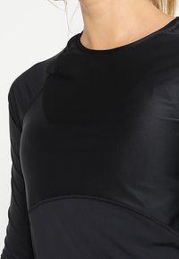 Schwarzes, athletisches Langarmshirt aus glattem, dehnbarem Stoff. Es verfügt über einen Rundhalsausschnitt und ein kontrastierendes Paneeldesign über der Brust.