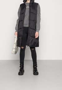 Svart pufferväst med hög krage, kombinerad med en grå sweatshirt, trasiga svarta jeans och kraftiga svarta stövlar. Håller en texturerad väska med ormskinnsmönster.