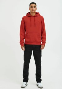 Red hoodie with a kangaroo pocket, drawstring hood, and embroidered logo. Paired with black jeans and gray-and-white sneakers.