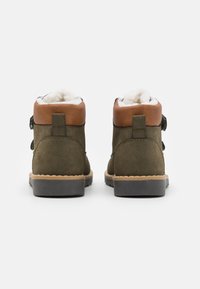 Bottines en daim vert avec un col en cuir marron et une doublure en laine blanche, comprenant une semelle texturée et deux sangles décoratives.