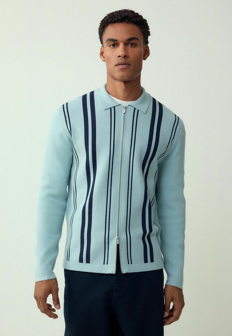 Hele sinine lukuga kardigan lahtise kraega ja tumesiniste vertikaalsete triipudega. Kangas näib sile ning kujundus on keha järgiv. Pikkade varrukatega.