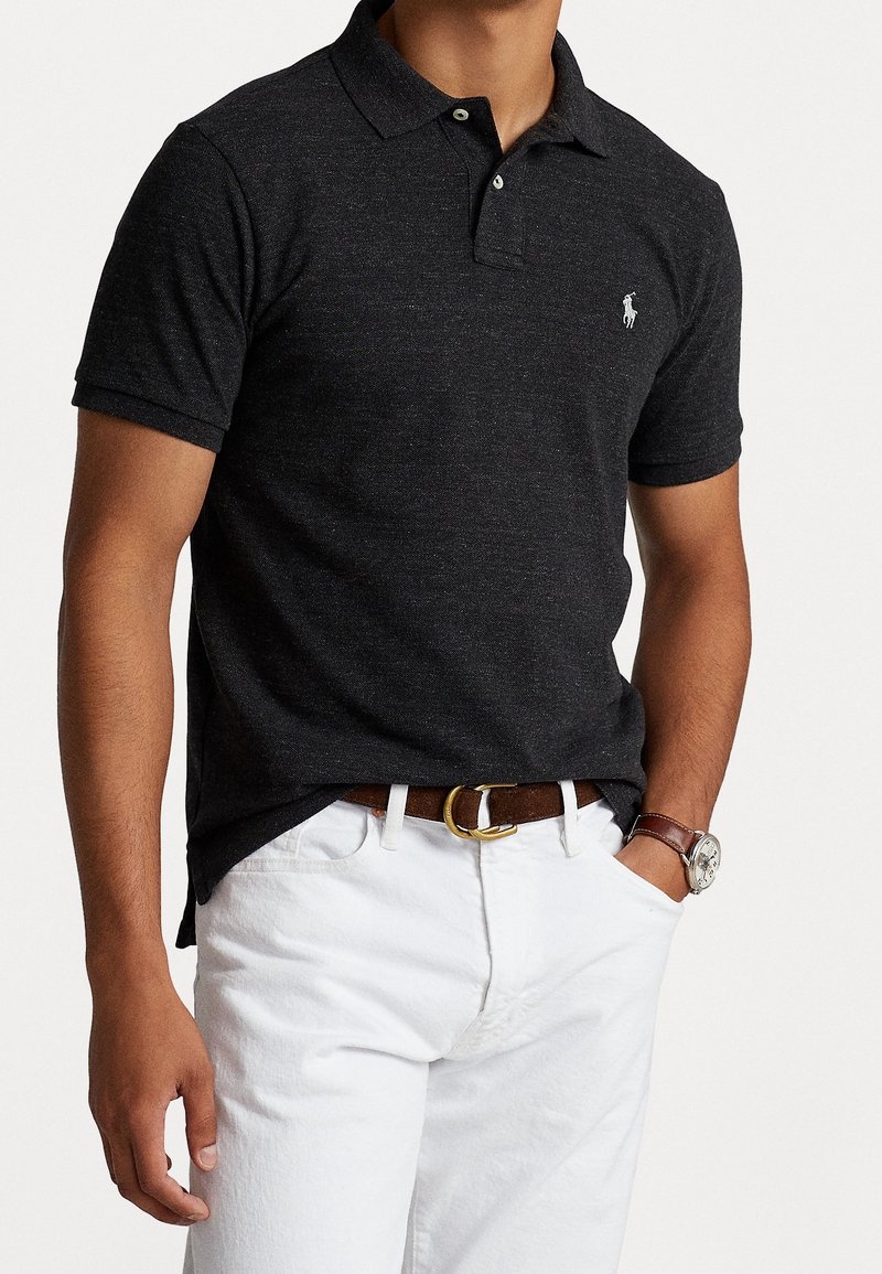 Homme portant un polo gris foncé à manches courtes avec un petit logo blanc, pantalon blanc, ceinture marron et montre-bracelet, main dans la poche.