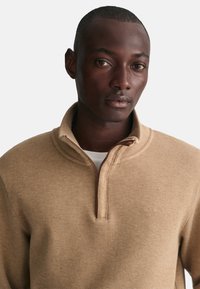 Beige ritsvest met een gebreid textuur, hoge kraag en subtiel logo. De stof lijkt zacht en warm, geschikt voor casual gebruik.