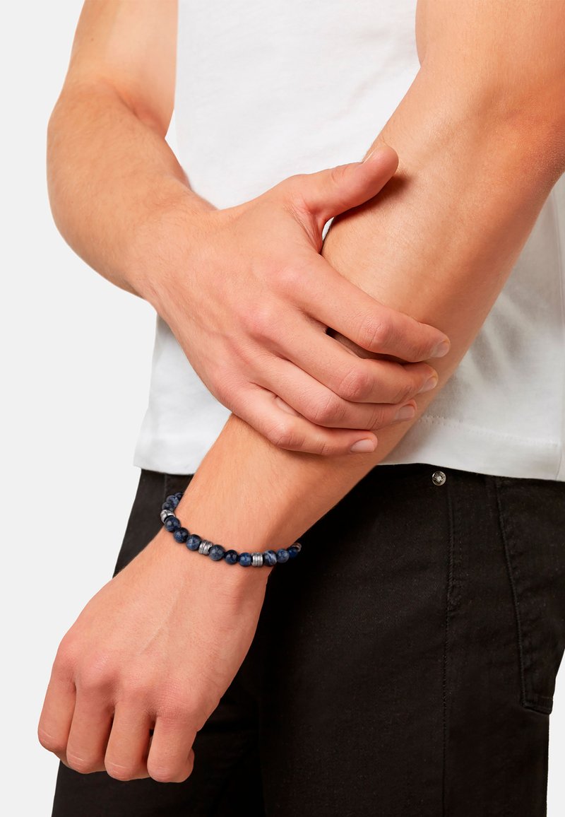 Hombre con una pulsera de cuentas azules sostiene su otro brazo, vestido con una camisa blanca y pantalones negros, frente a un fondo liso.