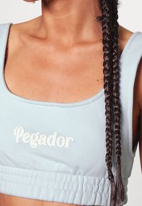 Jasnoniebieski krótki top z rozciągliwego materiału, z dekoltem w serek i haftem "Pegador" w białym kolorze. Włosy w warkoczu.