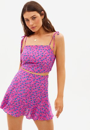 Jeune femme portant un ensemble deux pièces floral rose composé d'un crop top sans manches et d'une mini-jupe évasée sur fond blanc.