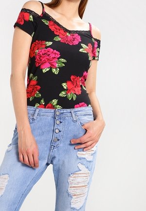 Vrouw draagt een zwarte cold-shoulder top met bloemenmotief in rood en roze, gecombineerd met lichtblauwe, hooggesloten jeans met scheuren.