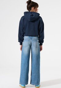 Femme de dos, portant un sweat à capuche bleu marine, un jean large bleu clair et des chaussures bleu clair avec des semelles en caoutchouc.