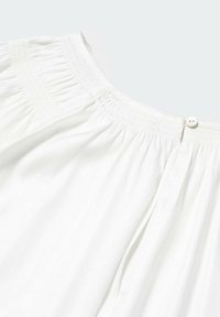 Tissu blanc avec détail smocké au niveau de l'encolure, fermeture par petit bouton et texture douce froncée.