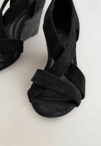 Sandales compensées noires en daim avec des lanières croisées, une texture douce, un bout rond et des détails tissés sur le talon compensé.