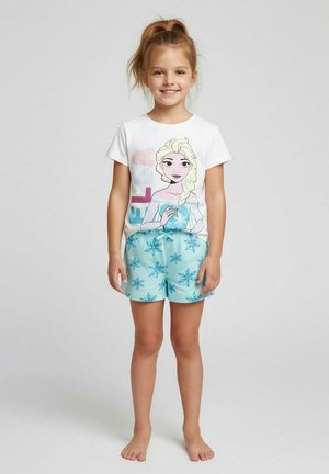 Junges Mädchen lächelnd, trägt ein weißes Hemd mit einer blonden, animierten Figur und hellblauen Shorts mit Schneeflockenmuster. Barfuß, neutraler Hintergrund.