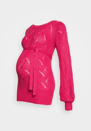 Pull de maternité en tricot rose avec un motif texturé, des manches longues et un lien à la taille, mettant en valeur le ventre.