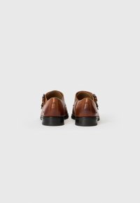 Zapatos de cuero marrón con un acabado suave, forma redondeada y suelas de goma negra. Presentan un detalle de hebilla lateral y costuras mínimas.