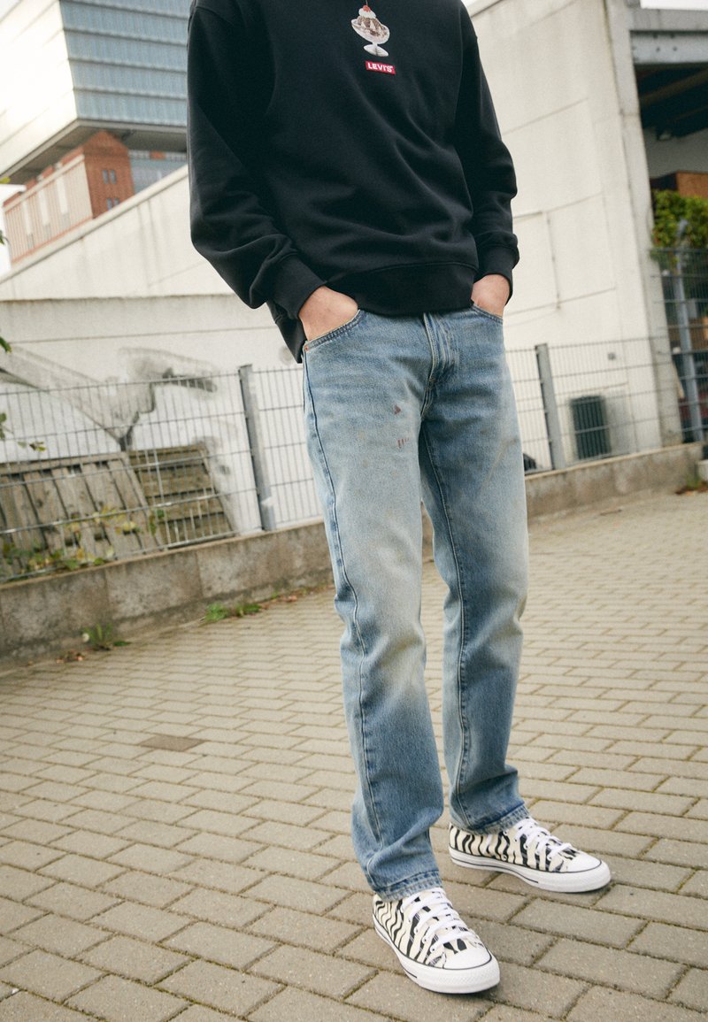 Jeans de mezclilla azul claro con un acabado desgastado y ligeros desgastes, combinados con unas zapatillas de lona a rayas blancas y negras y un sudadera negra.