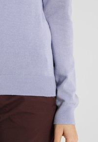 Jasnofioletowy sweter z kaszmiru z prążkowanymi mankietami i dołem. Dopasowany krój podkreśla miękką teksturę i gładkie wykończenie. W zestawie z ciemnobrązowymi spodniami.