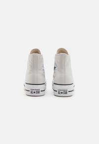Pár bílých vysokých tenisek Converse All Star zobrazených zezadu na jednobarevném bílém pozadí.