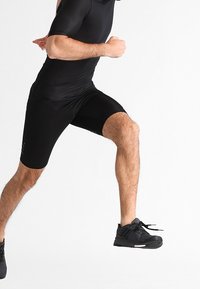 Bodysuit atlético negro con mangas cortas y pantalones cortos ajustados. Combinado con zapatillas negras texturizadas, demostrando movimiento.
