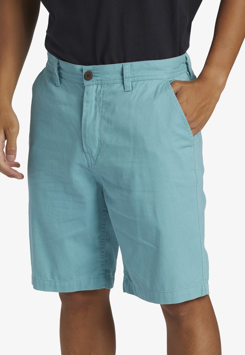 Ljus turkosa bomullsshorts med platt front, sidfickor och brun knappstängning i midjan. Slät textur och knälång längd.