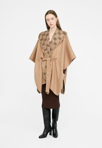 Poncho beige con colletto marrone a fantasia, legato in vita. Indossato sopra una gonna marrone e abbinato a stivali neri fino al ginocchio.