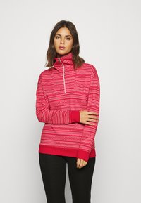 Rød fleece-pullover med høy krage, glidelås lukking og dekorative hvite mønstre. Har lange ermer og ensfargede mansjetter.