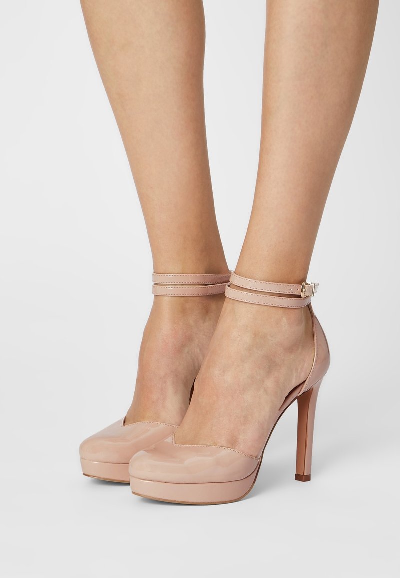 Jambes portant des escarpins en cuir verni couleur nude avec double bride à la cheville et bouts pointus sur un fond gris clair.