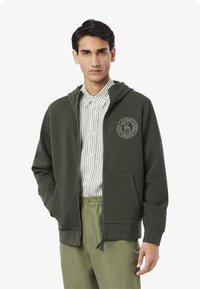 Felpa con zip verde oliva in tessuto testurizzato, con logo frontale e tasche, indossata sopra una camicia bianca a righe e pantaloni verde chiaro.