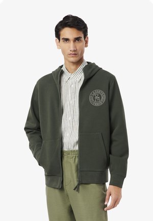 Felpa con zip verde oliva in tessuto testurizzato, con logo frontale e tasche, indossata sopra una camicia bianca a righe e pantaloni verde chiaro.