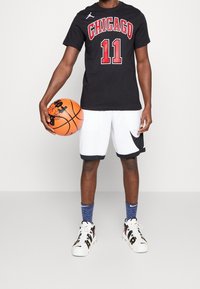 Camiseta negra con "CHICAGO" y el número "11" en rojo, combinada con pantalones cortos blancos con detalles en negro, calcetas azules con estampado y balón de baloncesto.