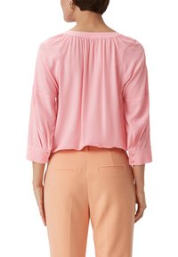 Helles rosa Bluse mit Dreiviertelärmeln, die eine geraffte Passe und Knopfakzente an den Ärmeln aufweist. Kombiniert mit hellen Pfirsich-Hosen.