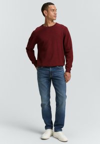 Pulover tricotat roșu cu guler rotund, asortat cu blugi albaștri și adidași albi. Puloverul are un model texturat.