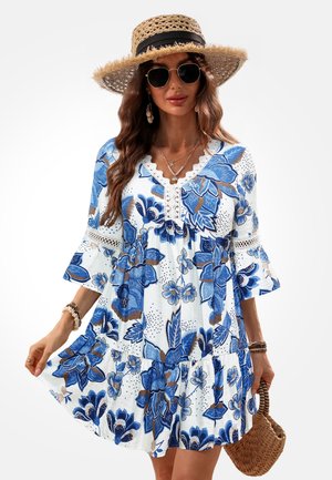 Femme portant un chapeau de paille à large bord, des lunettes de soleil et une robe blanche à motifs floraux bleus, tenant un sac tissé.