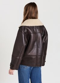 Giacca in pelle marrone con colletto in shearling beige, caratterizzata da una forma boxy, polsini con bottoni e dettagli di cucitura ben visibili. Indossata con jeans blu.