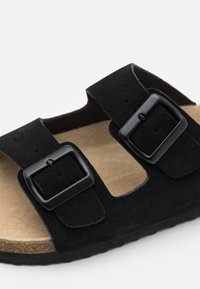 Juodos suede šlepetės su dviem reguliuojamais dirželiais su juodais sagtelėmis, bežinė paminkštinta pėdos dalis ir tekstūruota juoda išorinė dalis.