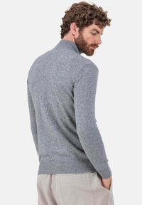 Pull gris en maille avec col montant et poignets côtelés, présentant une finition texturée et un matériau doux. Porté avec un pantalon de couleur neutre.