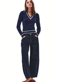 Suéter de punto estilo marinero de color azul marino con escote en V y dobladillo a rayas, combinado con unos jeans oscuros de corte holgado. El modelo está de pie con las manos en los bolsillos, luciendo zapatos rojos.