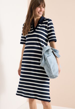Femme souriante, portant une robe à manches courtes rayée bleu marine et blanc, tenant un sac fourre-tout en denim bleu clair sur un fond neutre.