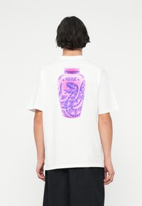 Valge puuvillane t-särk, mille seljal on suur lilla ja roosa purgi graafika, millel on madu ja tekst "NIKE". Mugav lõige lühikeste varrukatega.