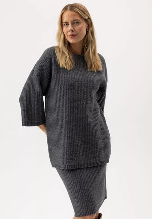 Donna bionda che indossa un maglione lavorato a maglia morbido color grigio scuro e una gonna aderente abbinata, in piedi su sfondo bianco.