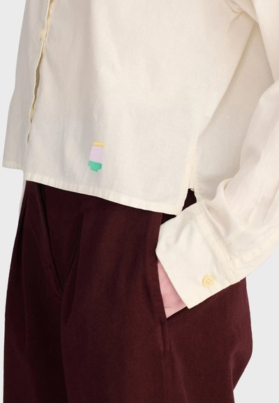 Personne portant une chemise crème boutonnée avec un petit patch multicolore et un pantalon marron foncé, main placée dans la poche du pantalon.