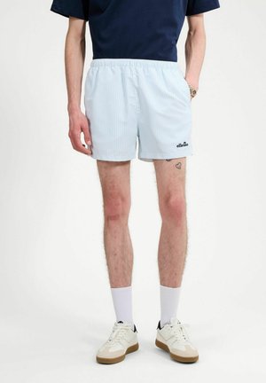 Mężczyzna noszący jasnoniebieskie, paskowane szorty z logo ellesse, białe skarpetki, białe trampki oraz granatową koszulkę, stojący z rękami w kieszeniach.