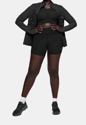 Žena stojí s rukama v bok, má na sobě černý sportovní podprsenku, odpovídající šortky s vysokým pasem, černou bundu, bílé ponožky a černé běžecké boty.