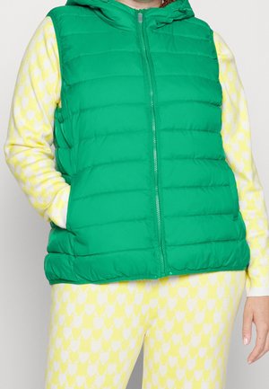 Person trägt eine grüne gesteppte ärmellose Weste über einem gelb-weiß gemusterten Langarmshirt und passenden Hosen.