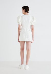 Λευκό μίνι φόρεμα με πλούσιες μανσέτες, υφή λουλουδιών, εφαρμοστή σιλουέτα και κλείσιμο με φερμουάρ πίσω, συνδυασμένο με λευκά πλατφόρμα παπούτσια.