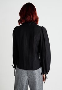 Blouse noire à côtes avec des manches bouffantes et des détails à nouer aux poignets. Associée à un pantalon rayé argenté brillant, conçu avec une coupe décontractée.