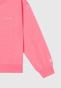 Sweatshirt rosa de manga longa com punhos canelados, logotipo "Champion" bordado parcialmente visível e pequeno emblema Champion perto do pulso.