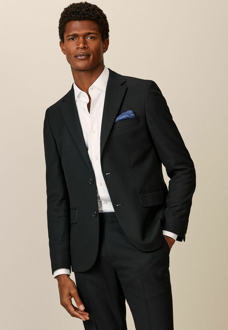 Traje negro con dos botones, camisa de dress blanca, pañuelo de bolsillo azul y corte a medida. Tejido suave y diseño estructurado.