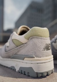 Light gray suede sneaker with a textured green mesh accent, featuring a white midsole and a gray rubber outsole. "NB" logo on heel.