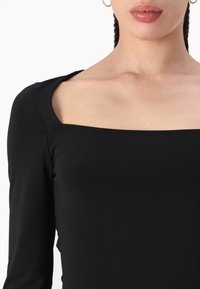 Juodas ilgomis rankovėmis bodis su kvadratiniu iškirpimu, pagamintas iš lygaus, tampraus audinio. Turi aptemptą siluetą ir minimaliai siuvinėjimo detalių.