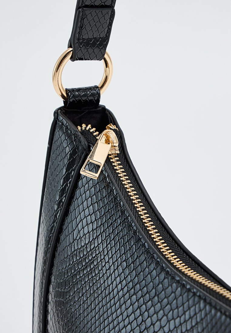 Sac à main noir texturé avec fermeture éclair dorée et attache de poignée ronde dorée, gros plan sur le détail de la fermeture éclair et de la sangle sur un fond clair.
